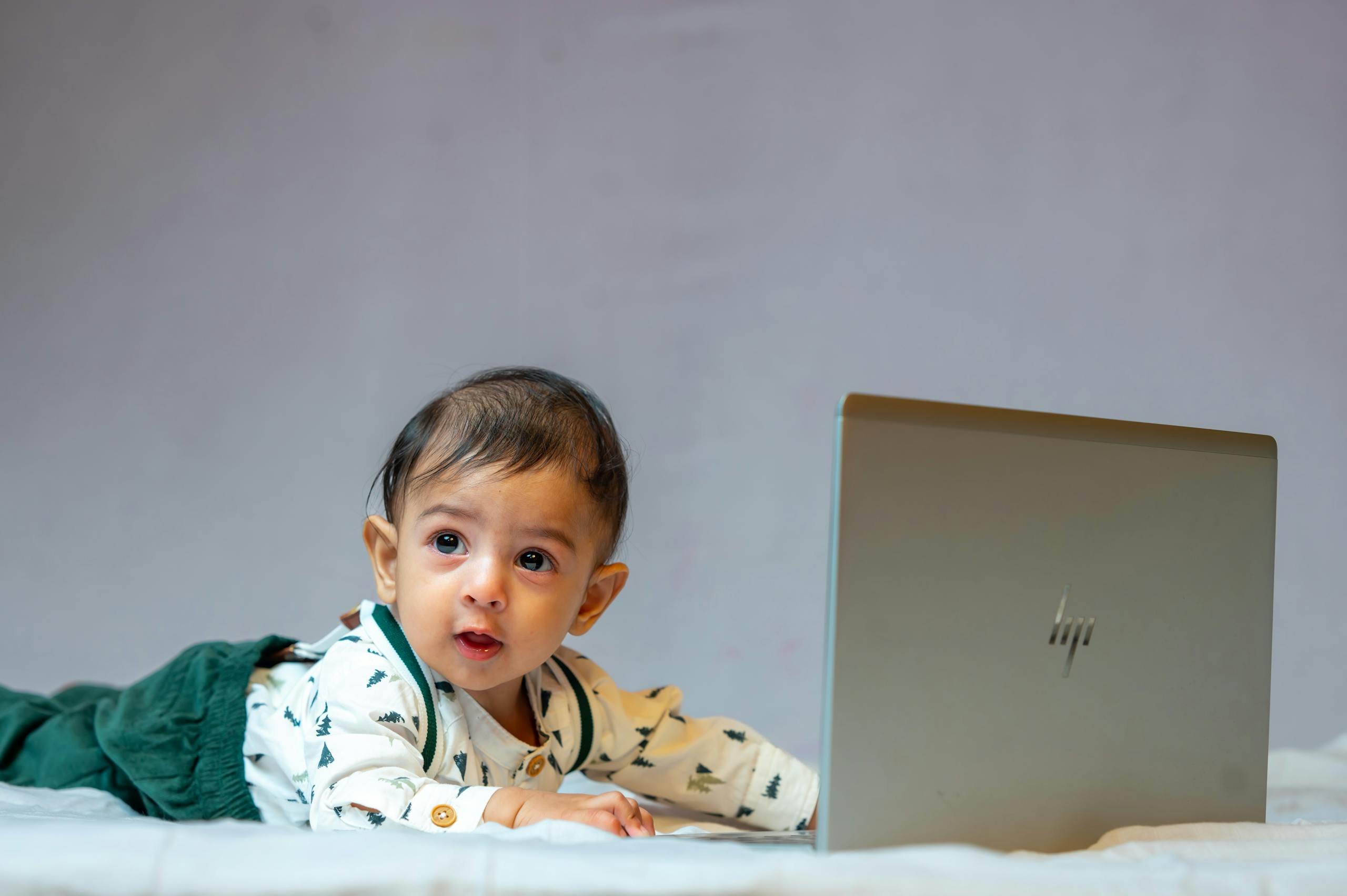 A cute baby lying on bed with a laptop, exploring technology indoors.
