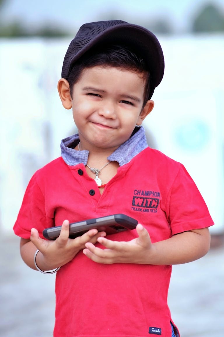 A cute South Asian boy in a red shirt smiling while holding a mobile phone outdoors.