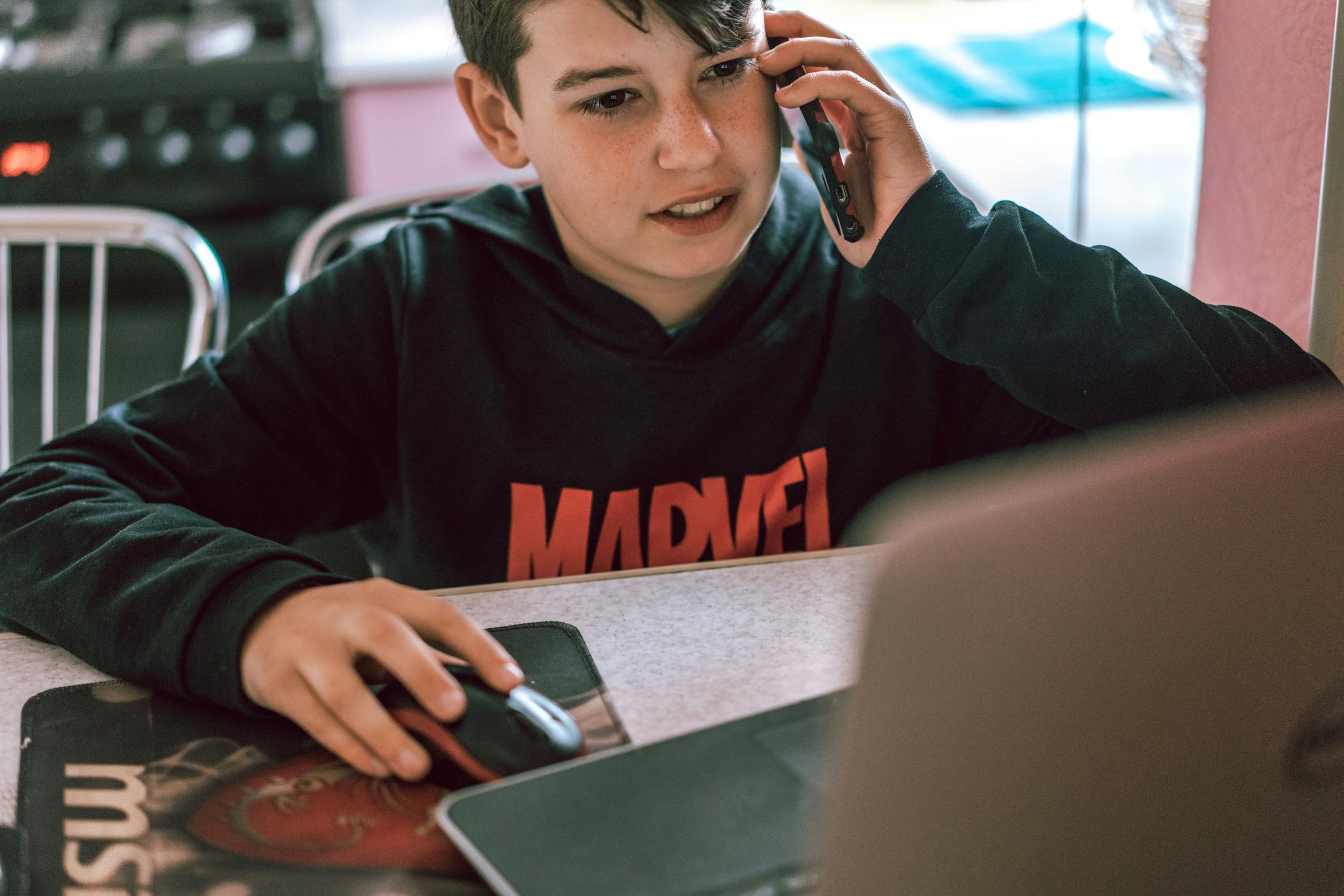 Teenage boy using a phone and laptop, engaging in multitasking indoors.